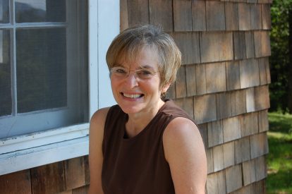 Picture of smiling woman with glasses wearing a brown sleeveless top with a background of a home with shingles