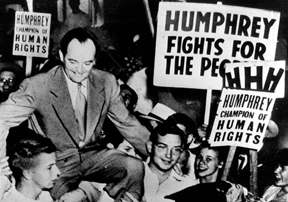 Black and white image of a man sitting on the shoulders of other men, with signs being held up around him with the text "HUMPHREY CHAMPION OF HUMAN RIGHTS" on them.