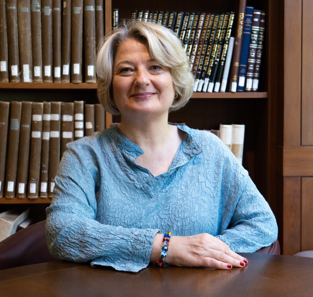 A smiling woman sits at a table in an indoor setting with a desk and a library in the background.