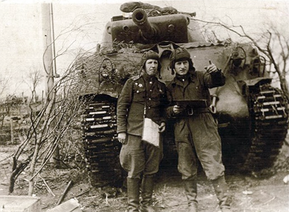 Sepia picture of two men in military gear in front of a WWII Tank